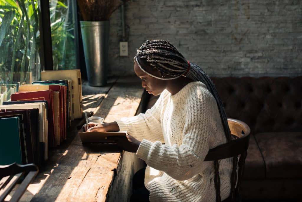 Woman with dreadlocks sitting in a sunlit room journaling at a table.
