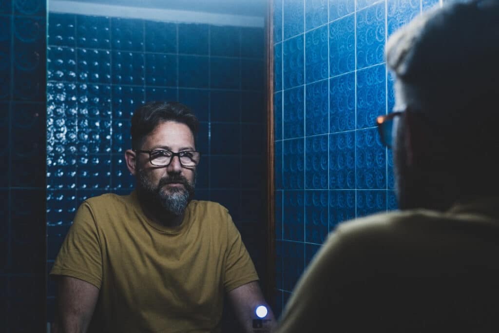 Man in a yellow shirt wearing glasses looks at himself in the mirror in a blue-tiled bathroom.